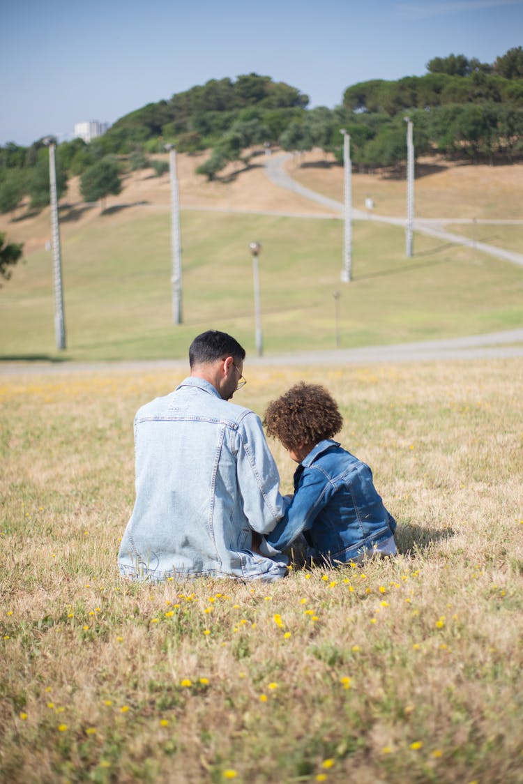 A Couple In Denim Jackets Sitting On Green Grass Field