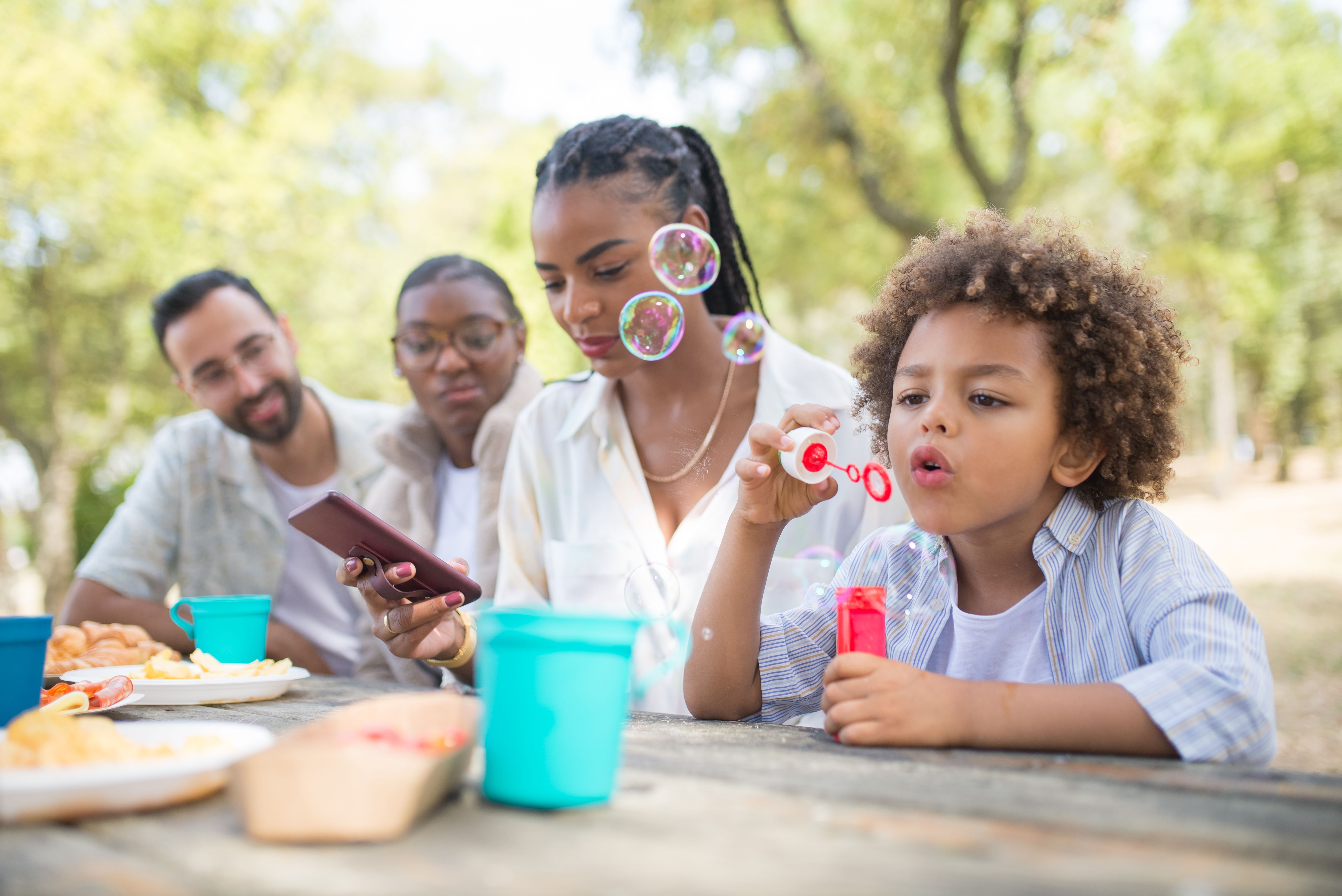A cheerful family enjoying a picnic outdoors, with a child blowing bubbles and adults engaging with smartphones.