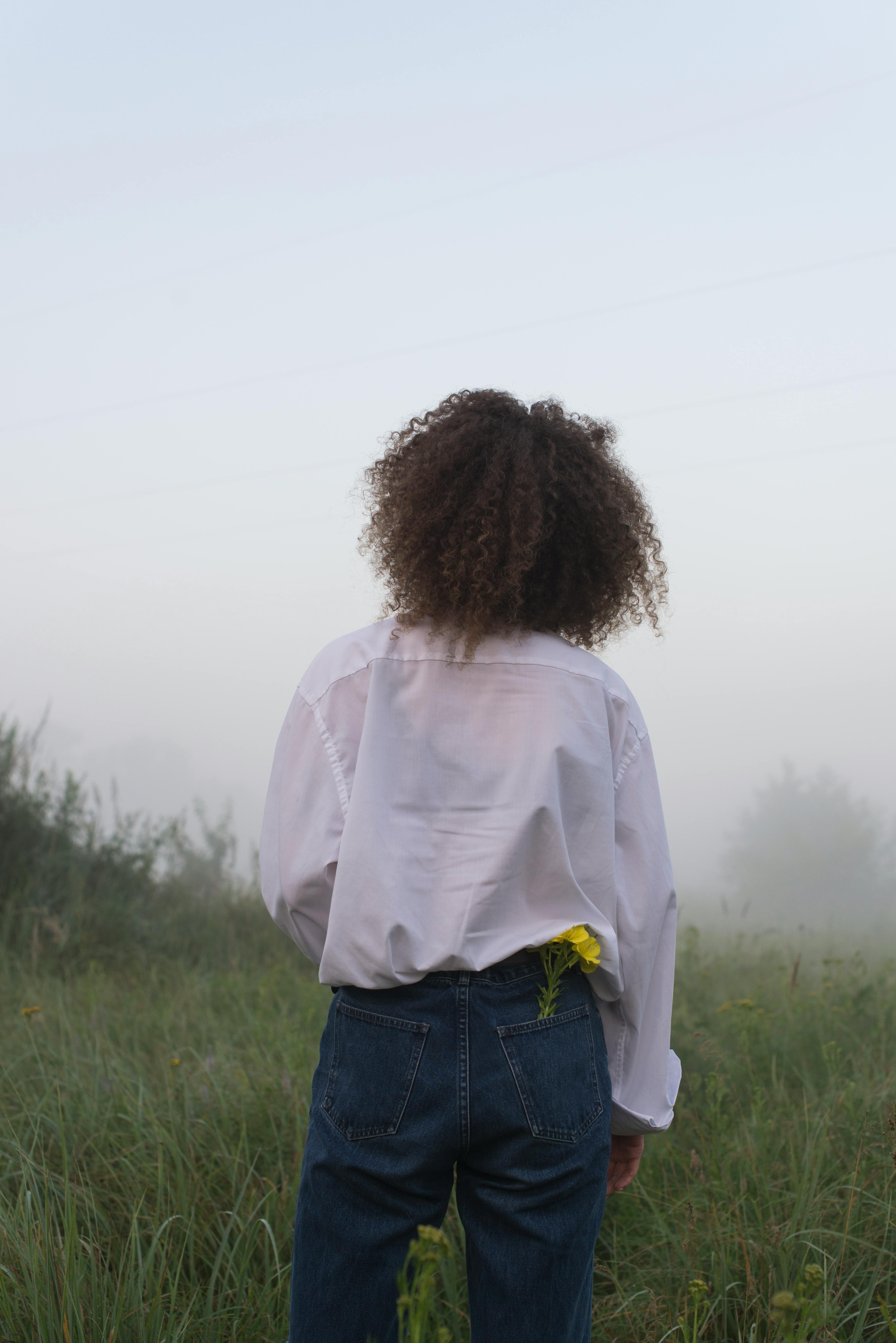 A woman with curly hair stands in a foggy field with a yellow flower tucked in her jeans pocket.