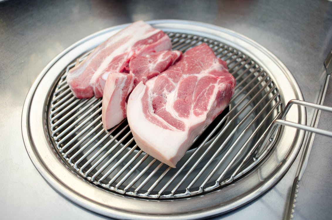The image shows raw pork cuts placed on a round metal grill, ready for cooking. The meat has visible layers of fat and muscle, showcasing its marbling. The grill sits within a stainless steel pan, suggesting preparation for a barbecue or grilling session. The setting reflects a simple and clean setup, emphasizing the fresh quality of the meat.