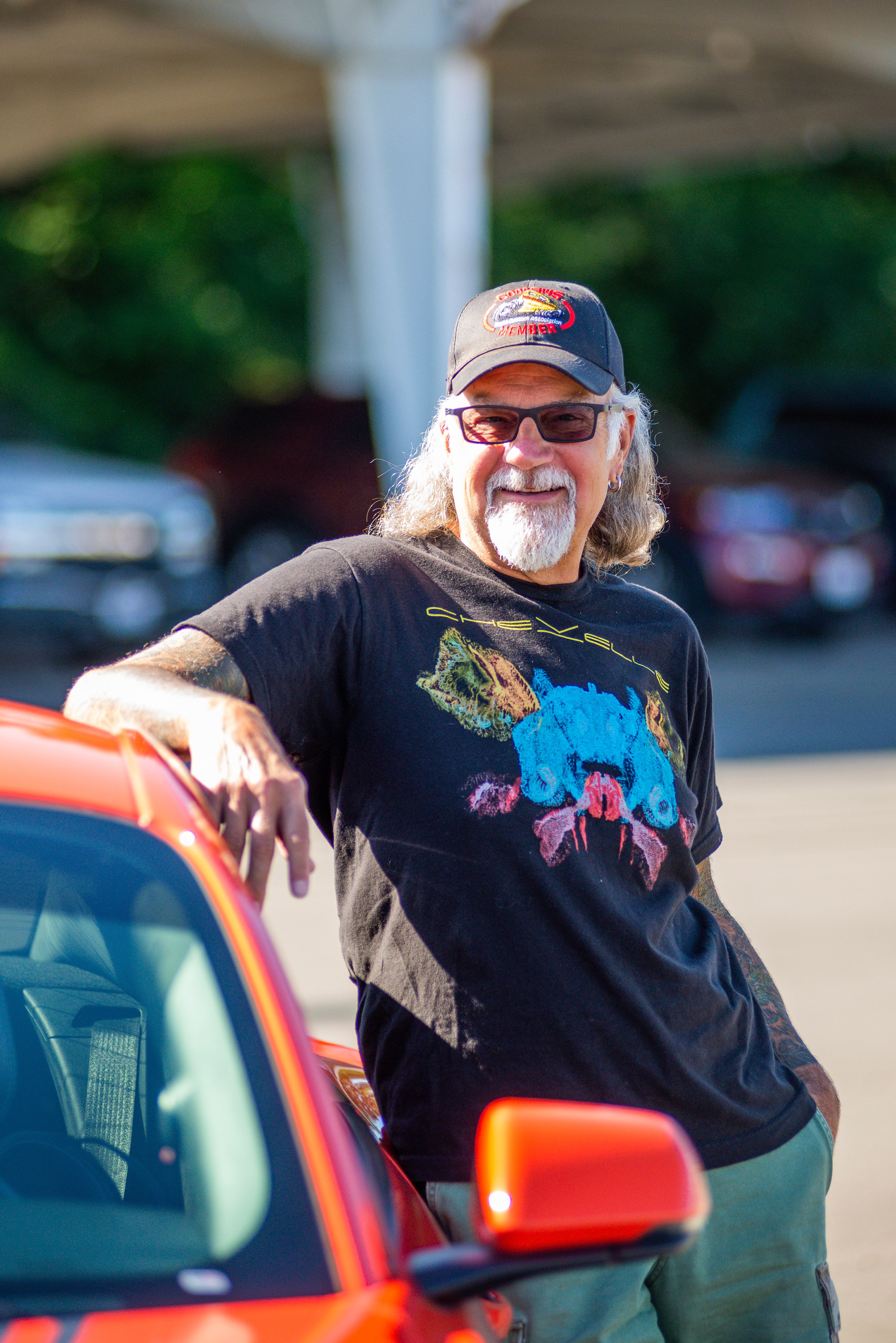 Elderly man with sunglasses and hat leaning on red car in sunlight.