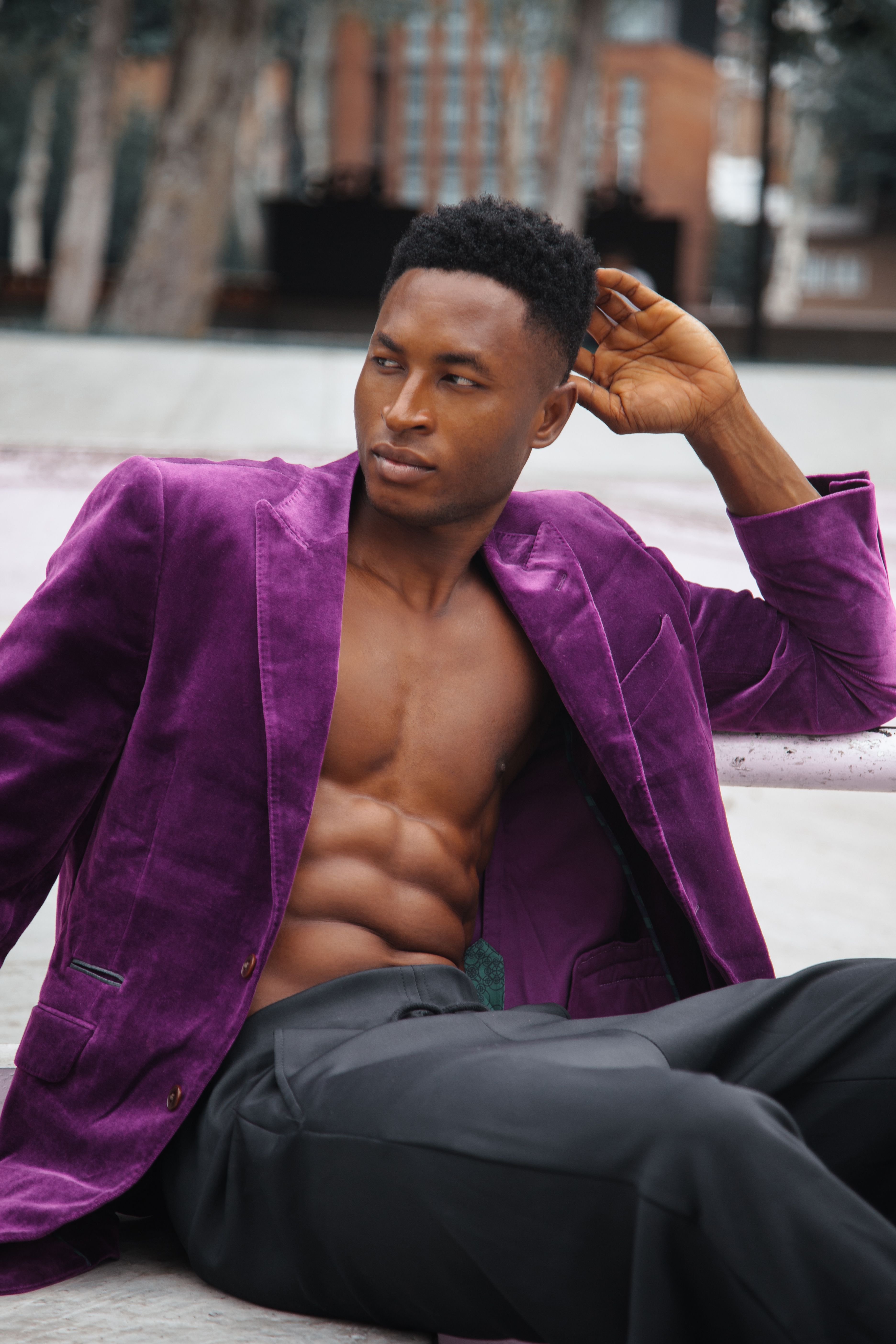 Fashionable man in purple blazer poses confidently on a bench in an outdoor setting.