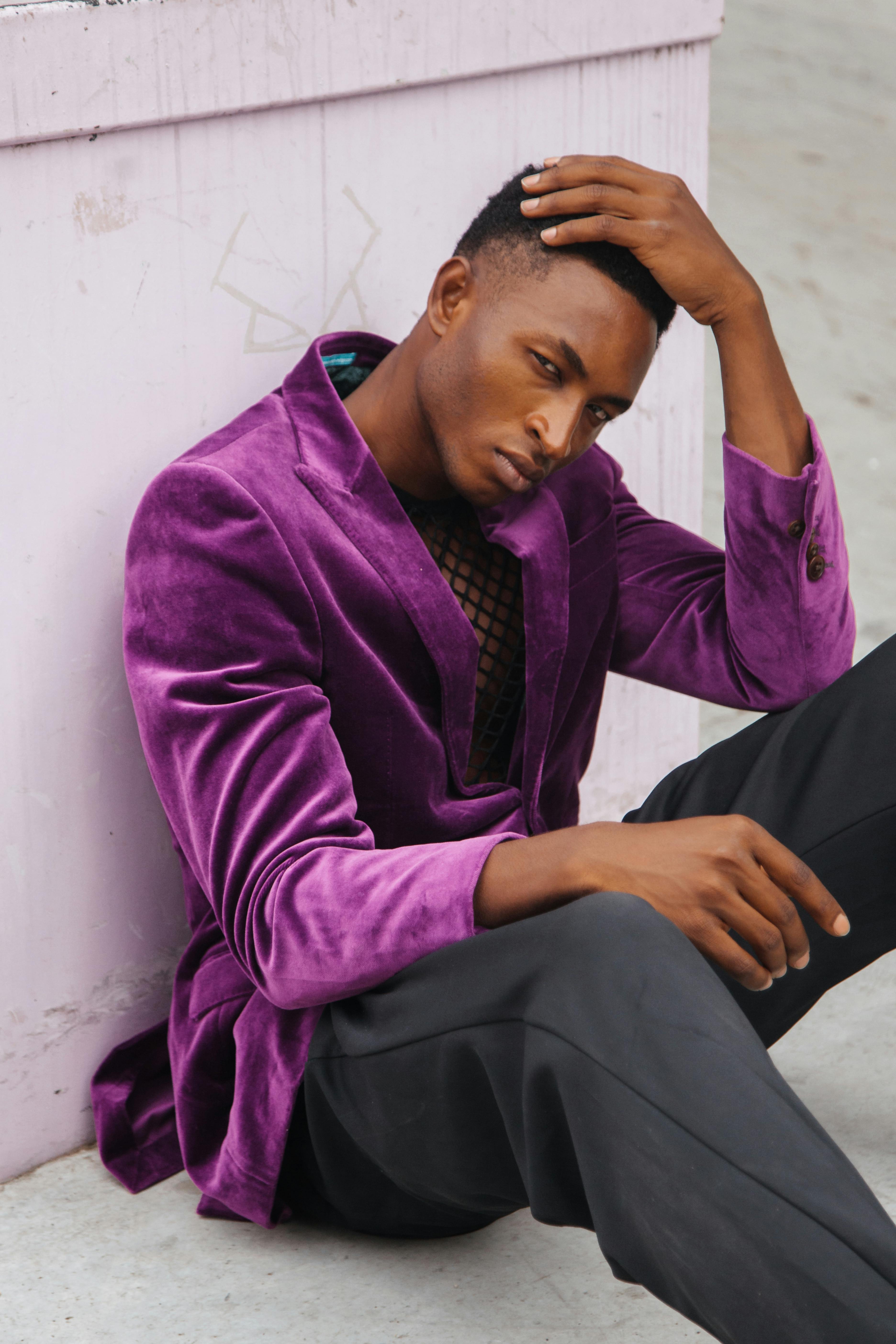 Stylish African American man in a purple jacket seated against a wall, showcasing modern menswear fashion.