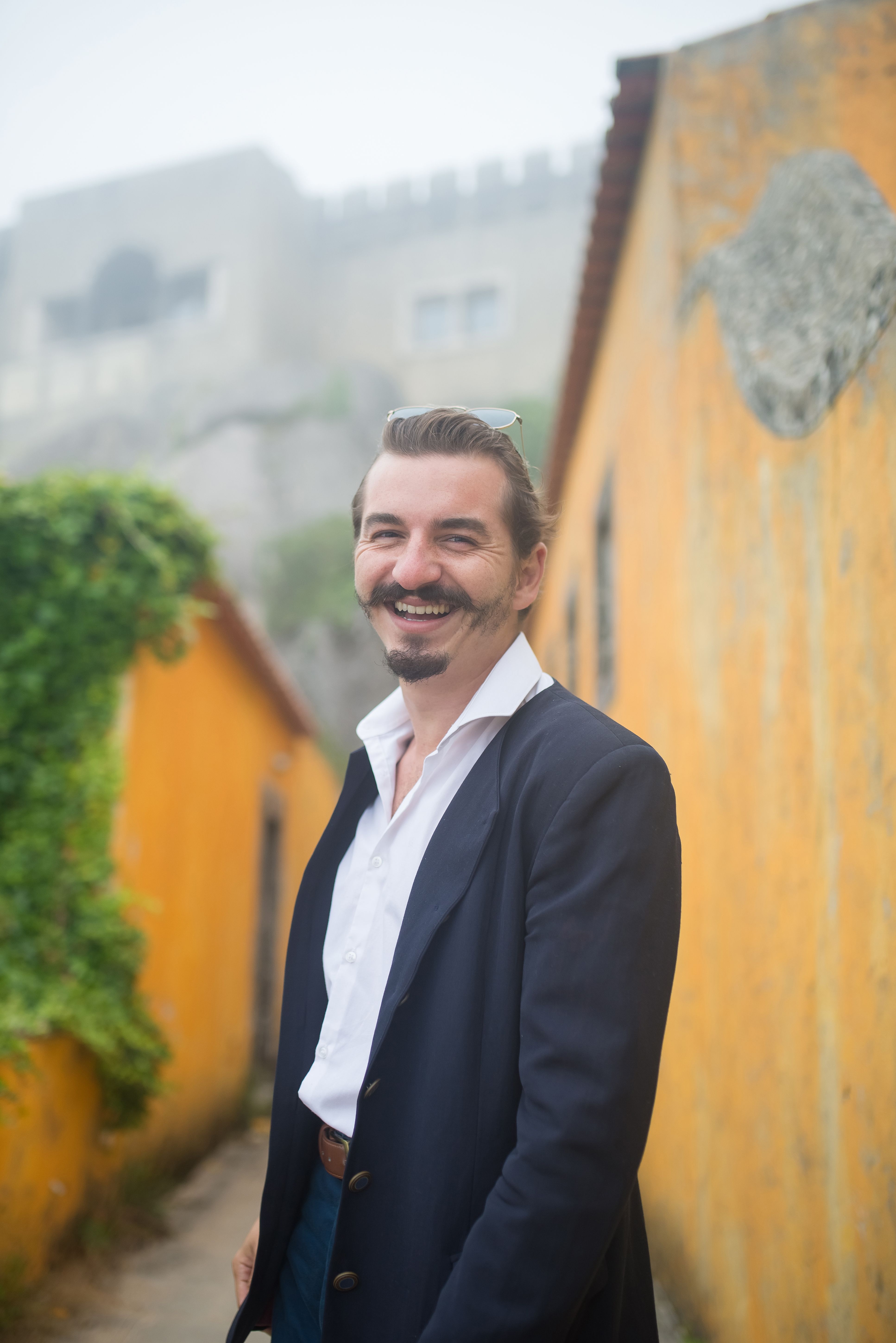 A cheerful man in a suit jacket stands in a vibrant, historic street, exuding confidence and joy.