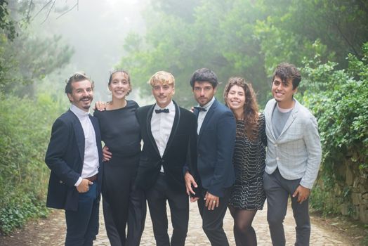 A cheerful group of young adults in formal wear posing on a foggy, tree-lined path.