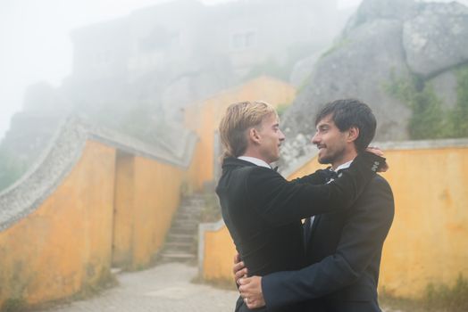 Gay couple in suits embracing on their wedding day, with a misty, romantic backdrop.