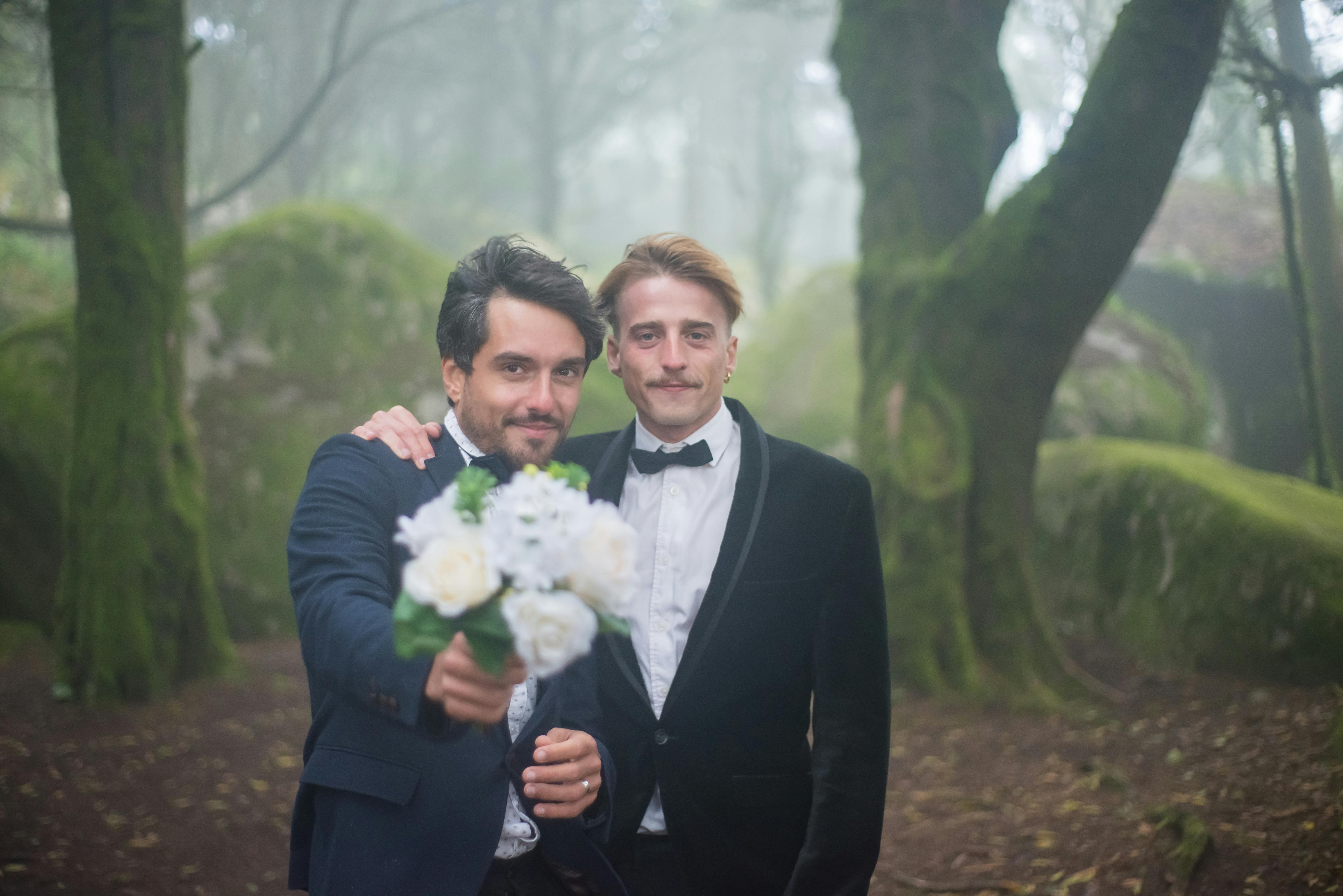 a man in black suit standing beside his partner holding a bouquet of flowers