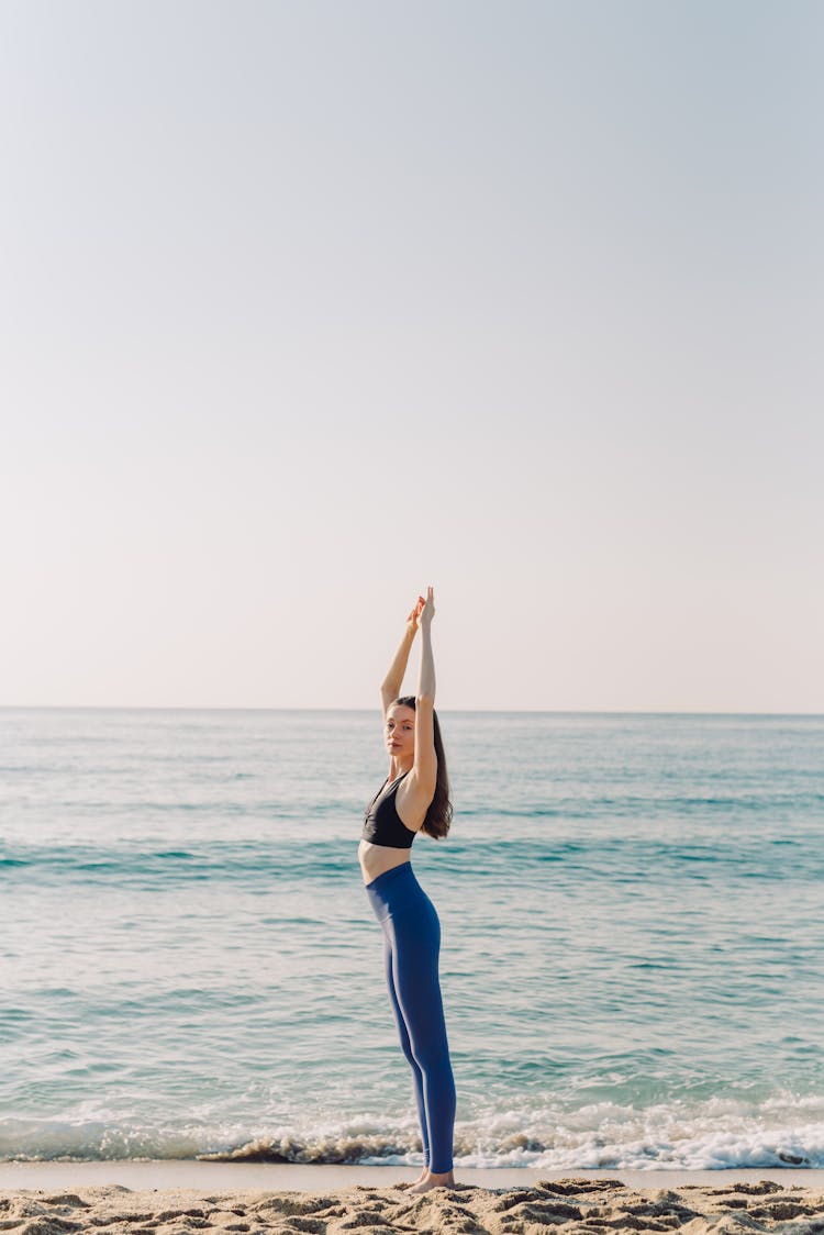 Fit Woman In Blue Leggings And Black Sports Bra Doing Yoga On Sea Shore 