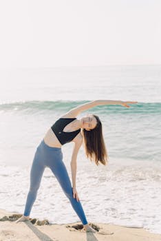 A woman in activewear performing yoga stretches on a sandy beach by the sea.