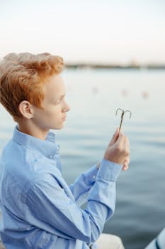Young boy with red hair examining a fishing hook by a calm lakeside during the day.