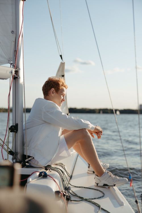 Free A young boy in white attire sits on a sailboat, gazing at the ocean during summer. Stock Photo