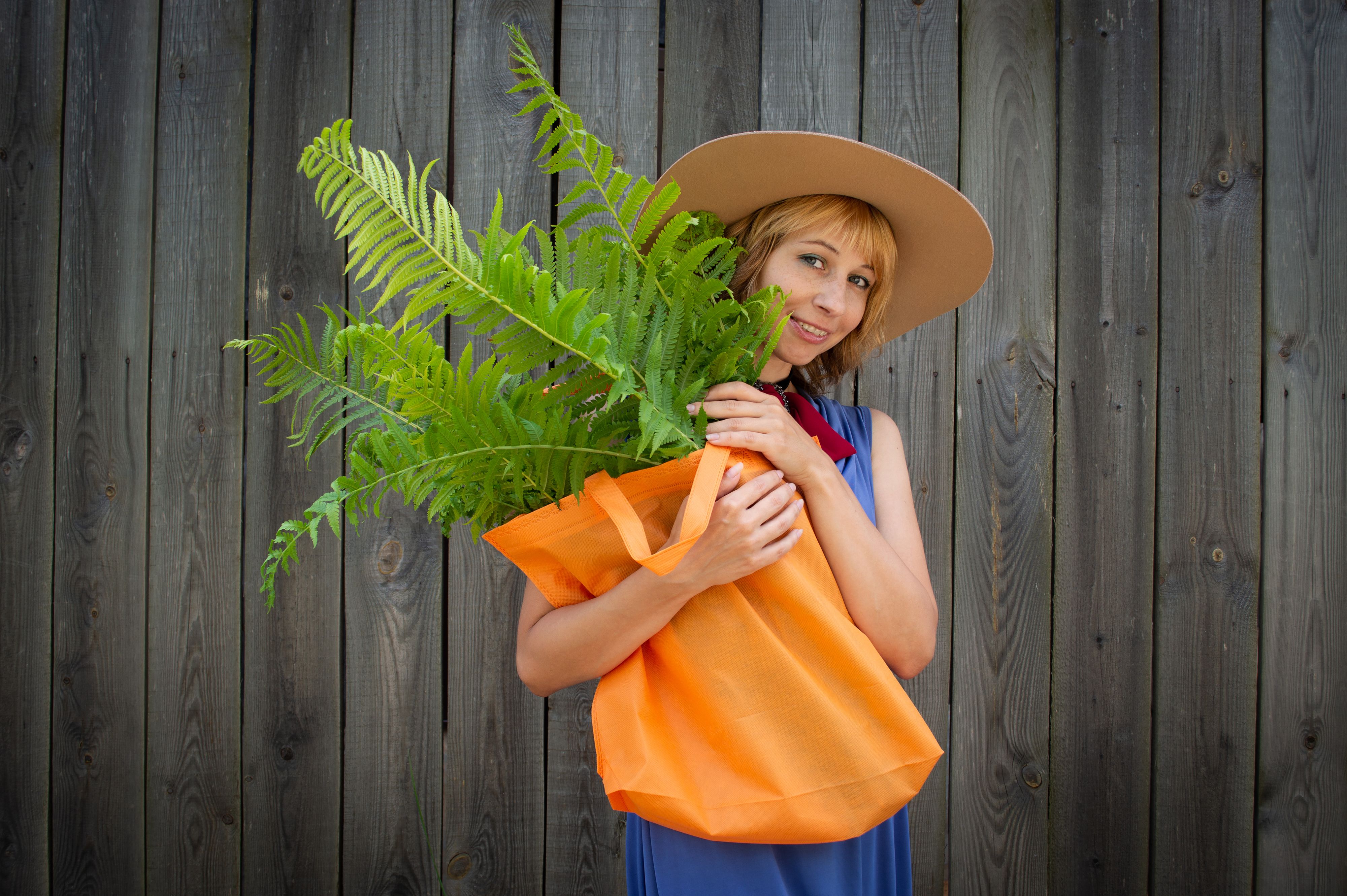 A woman in a hat holding an eco-friendly bag with ferns against a wooden backdrop.