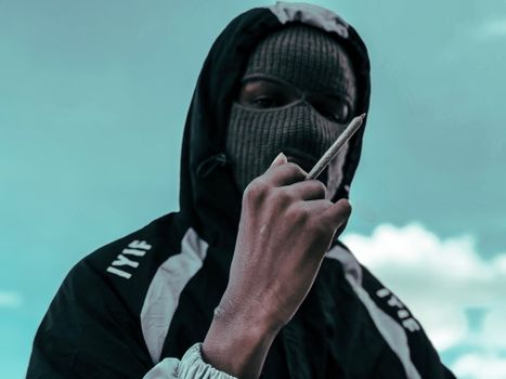 A person wearing a full face mask holds a rolled cigarette in an outdoor setting against a cloudy sky.