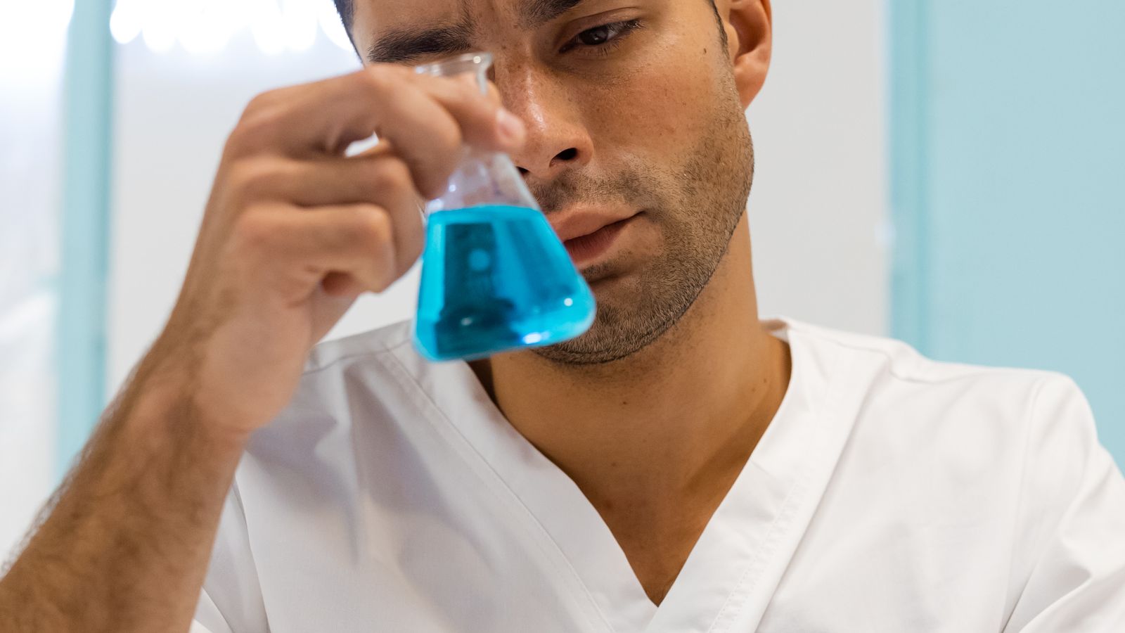 Scientist in lab coat examining blue liquid in flask, focused on experiment.