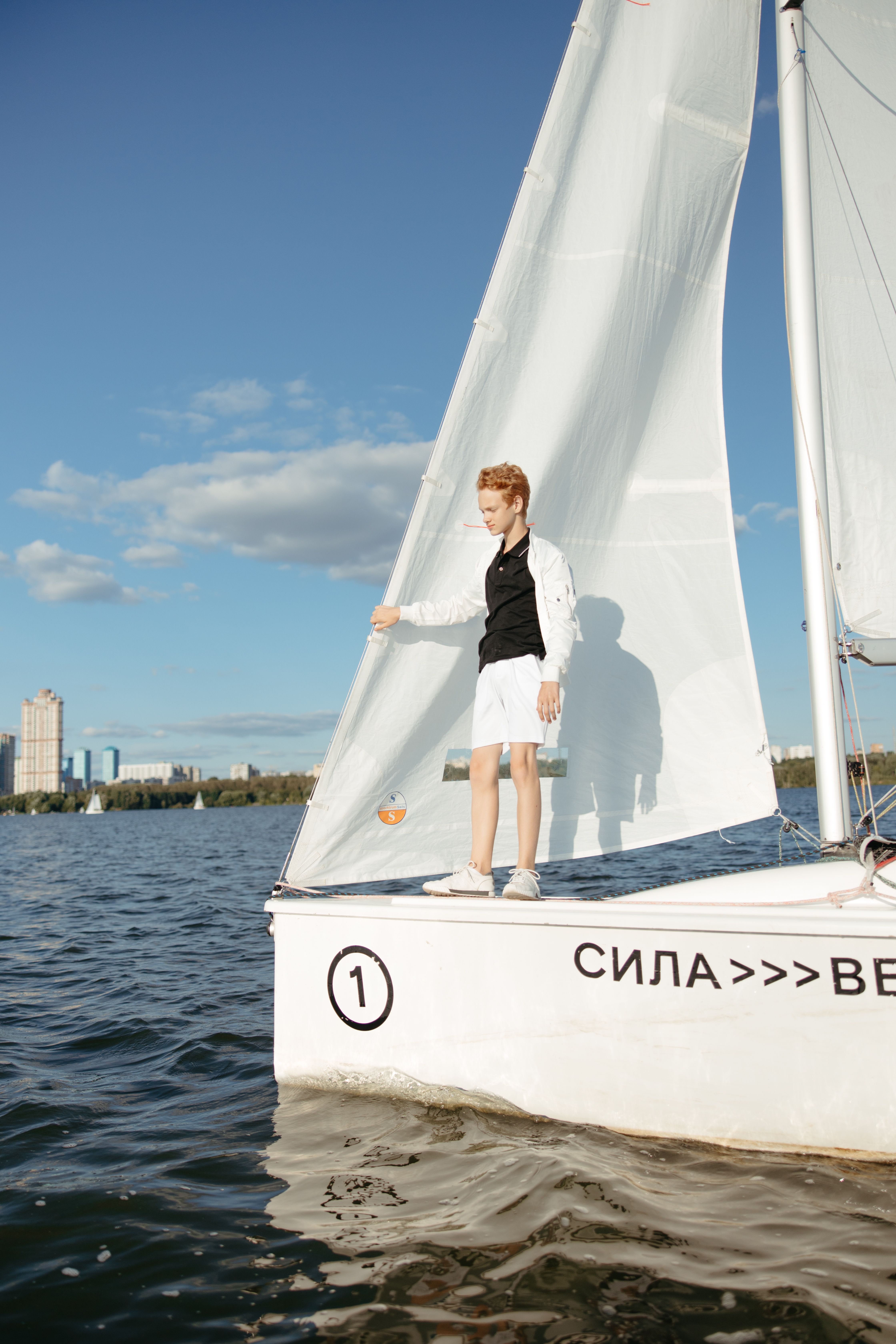 A young boy enjoys sailing on a clear day, standing on a sailboat in bright sunlight.