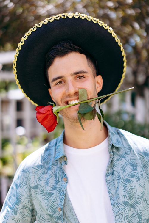 Free Portrait of a smiling young man wearing a sombrero and holding a red rose in his mouth. Stock Photo