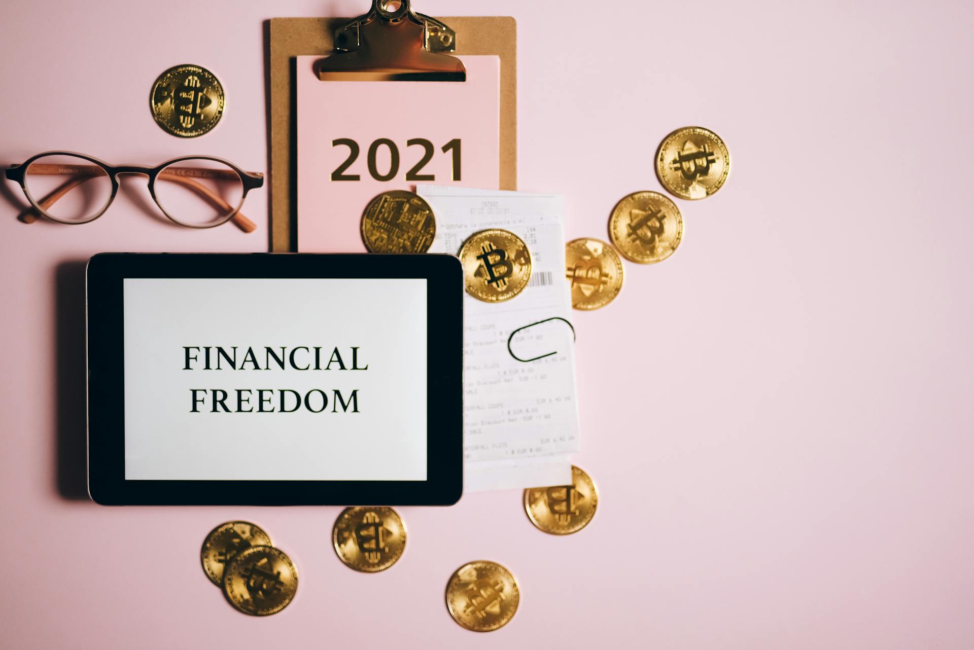 Flat lay image of financial freedom concept with bitcoins, tablet, and clipboard on a pink background.