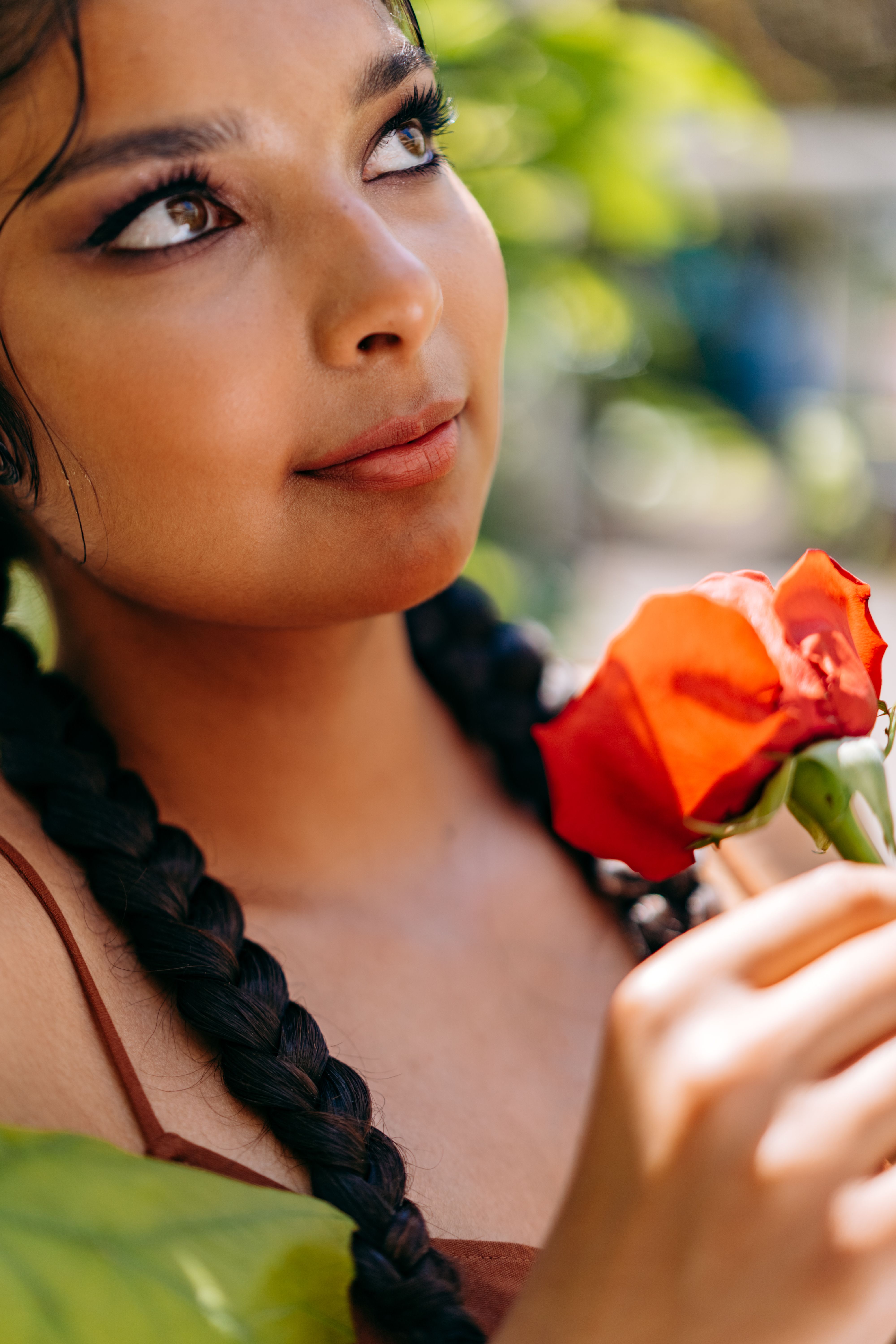 Portrait of a woman with braided hair holding a red flower, set against a lush outdoor background.