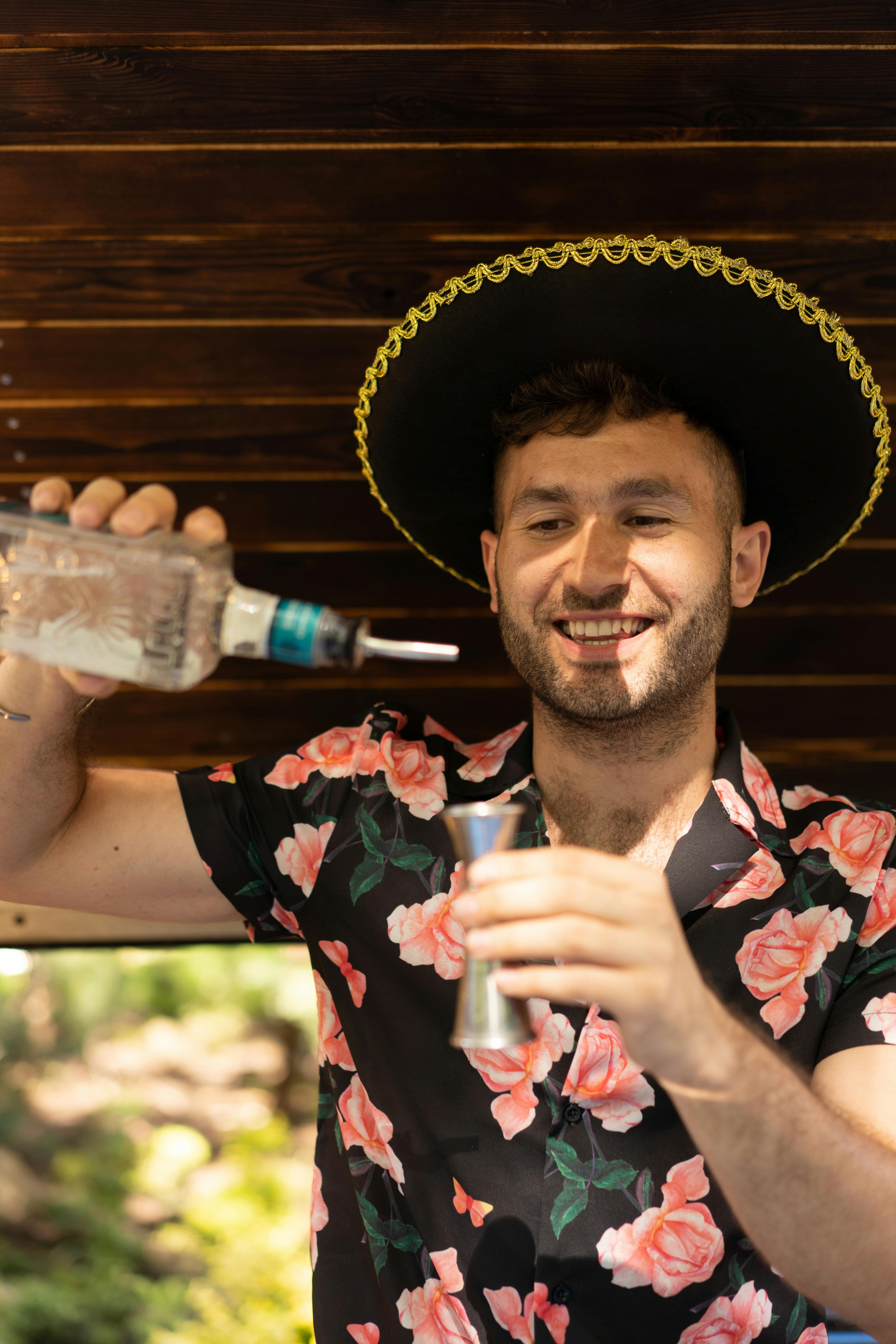 Happy bartender wearing a sombrero pours tequila into a jigger outdoors.