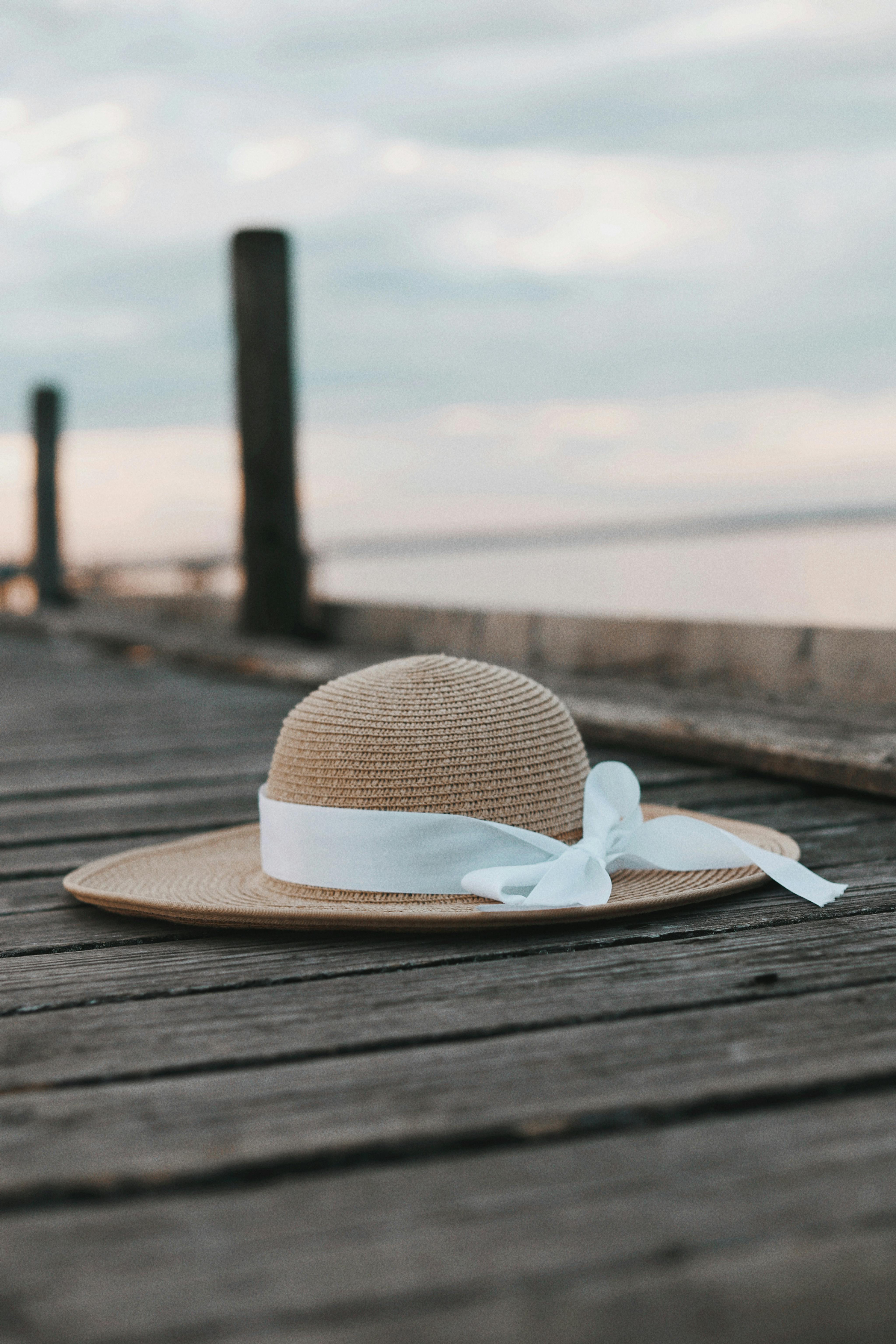 A straw hat adorned with a white ribbon rests on a wooden pier by the water.