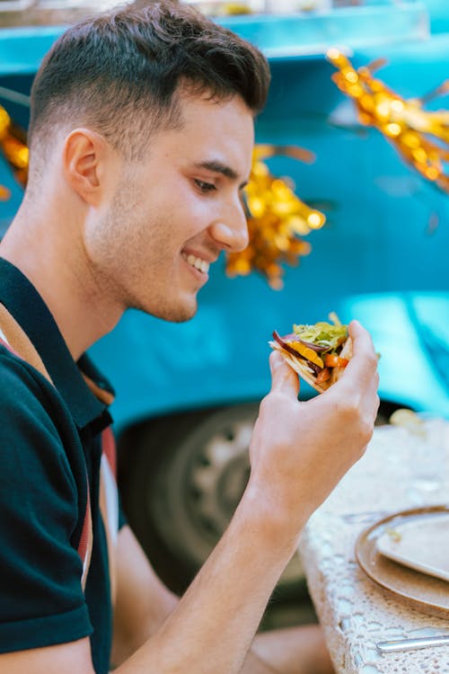 Free A young man smiling as he enjoys a delicious taco outdoors with festive decor in the background. Stock Photo