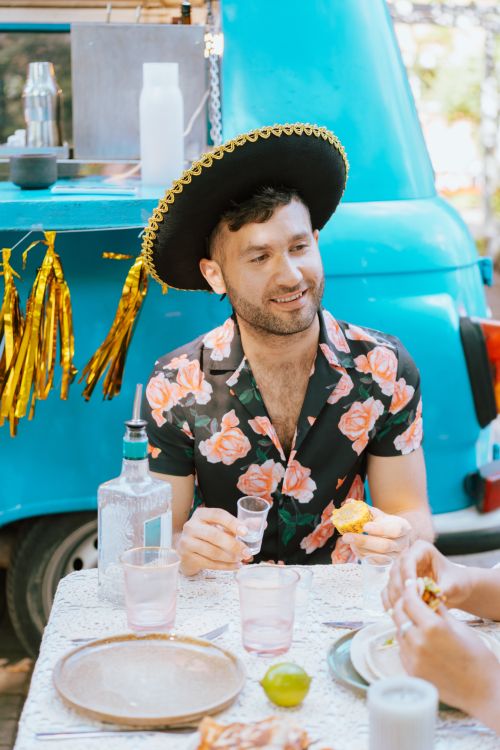 Free Man in a floral shirt and hat enjoying a meal outdoors by a blue food truck. Stock Photo