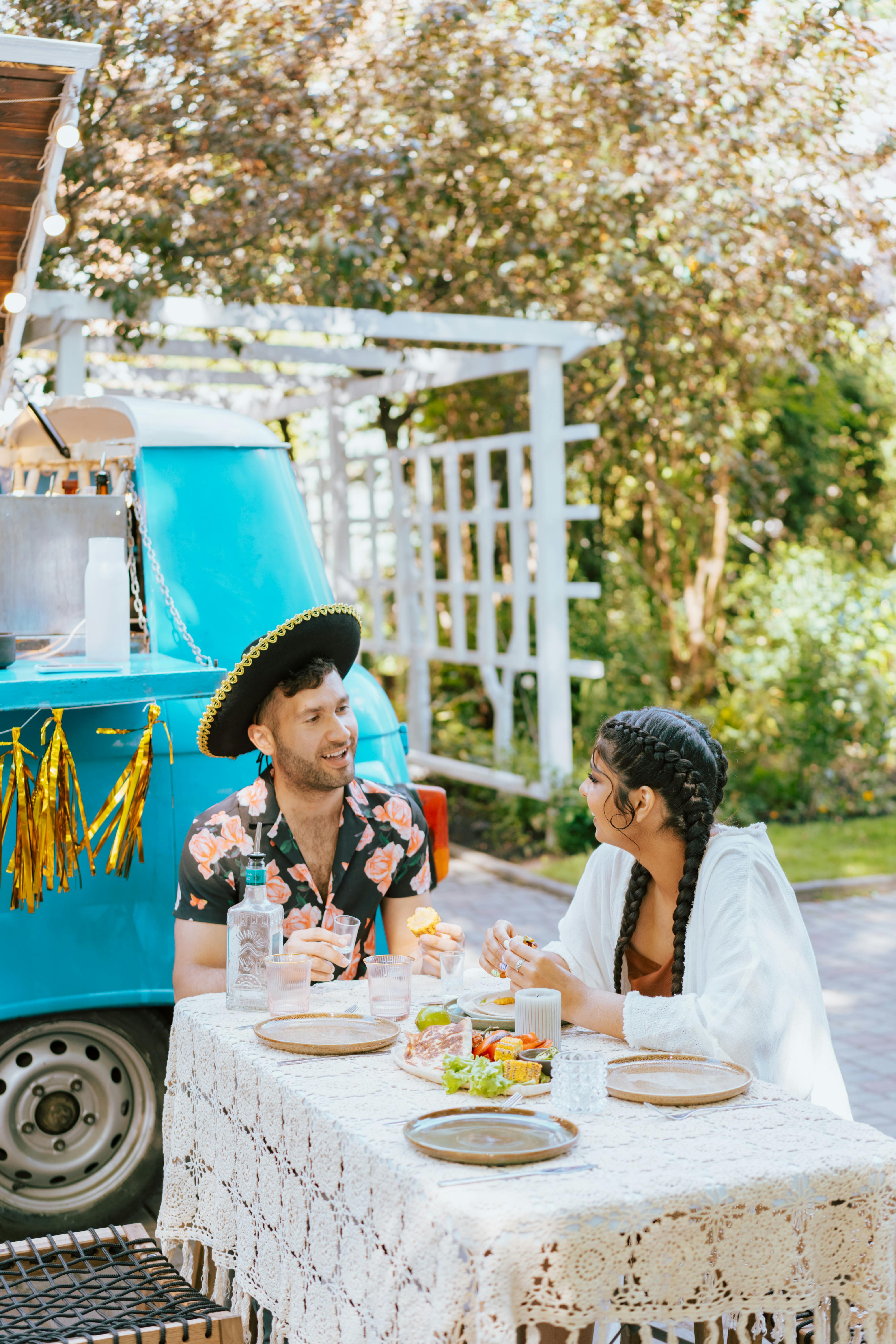 A couple enjoys Mexican food and drinks at an outdoor table beside a vibrant blue food truck.