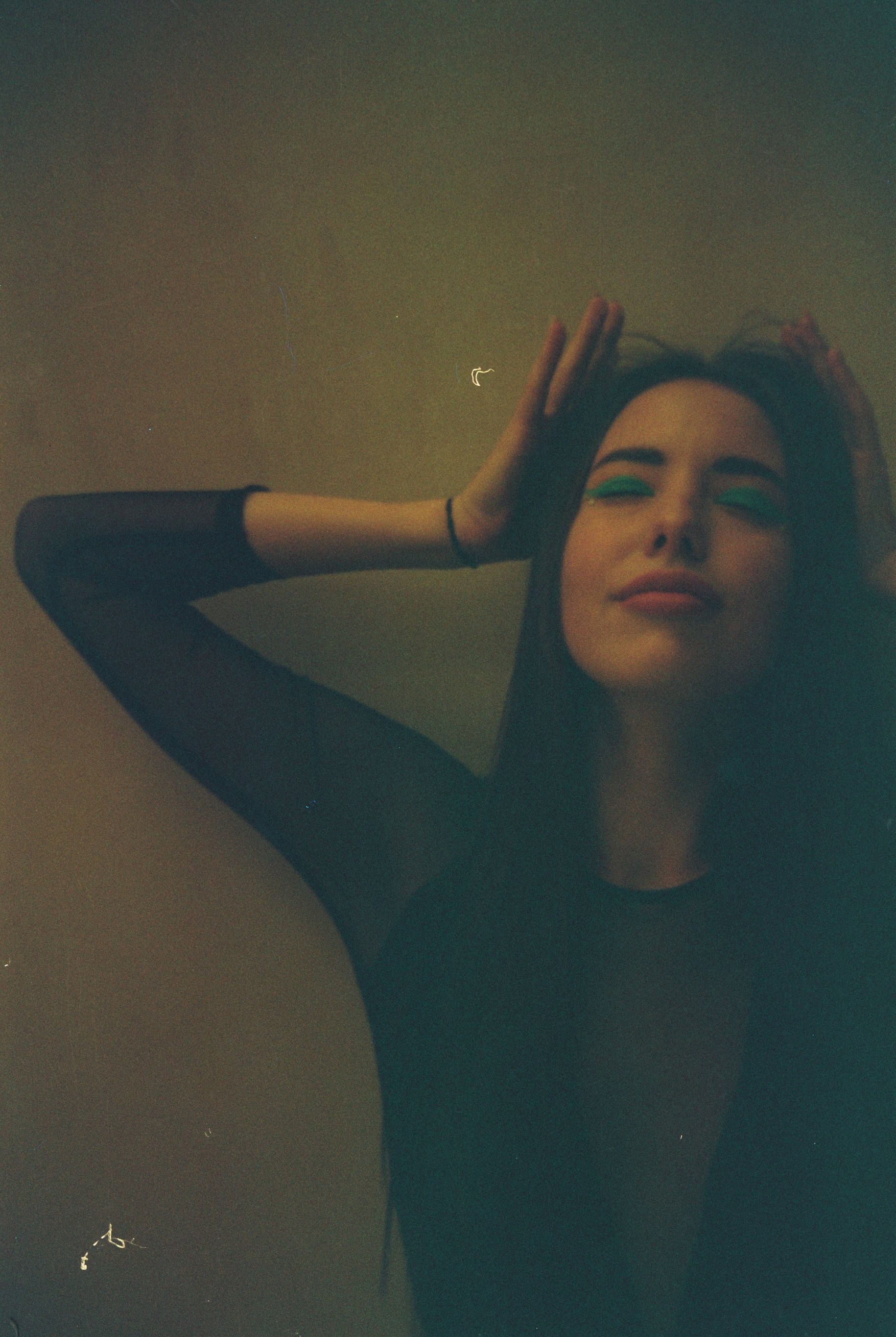 A moody portrait of a woman indoors, her eyes closed with hands on head.
