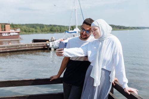 Free Happy Muslim couple taking a selfie by the lake on a sunny day, capturing joyful moments together. Stock Photo