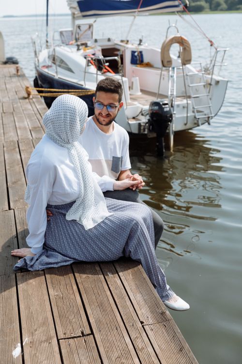 Free A young couple sitting on a wooden dock beside a sailboat, enjoying a sunny day. Stock Photo