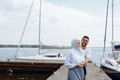 Free Joyful couple laughing together on a wooden dock by the marina, surrounded by boats and clear skies. Stock Photo