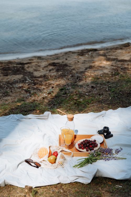 Free A picturesque picnic by the seashore with fruits, drinks, and a wooden tray on a blanket. Stock Photo
