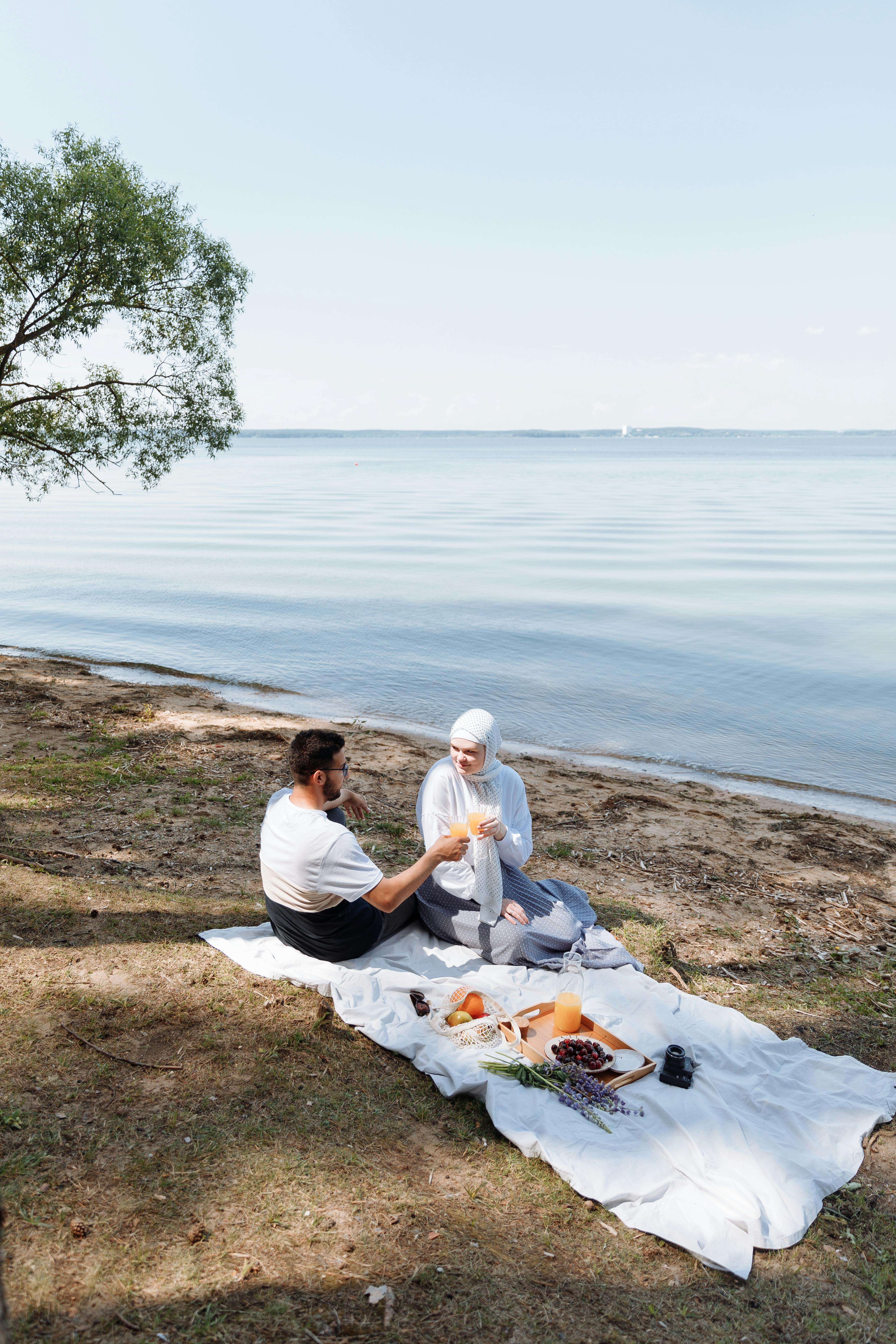 A couple enjoying a romantic picnic by the lake with fruits and drinks on a sunny day.