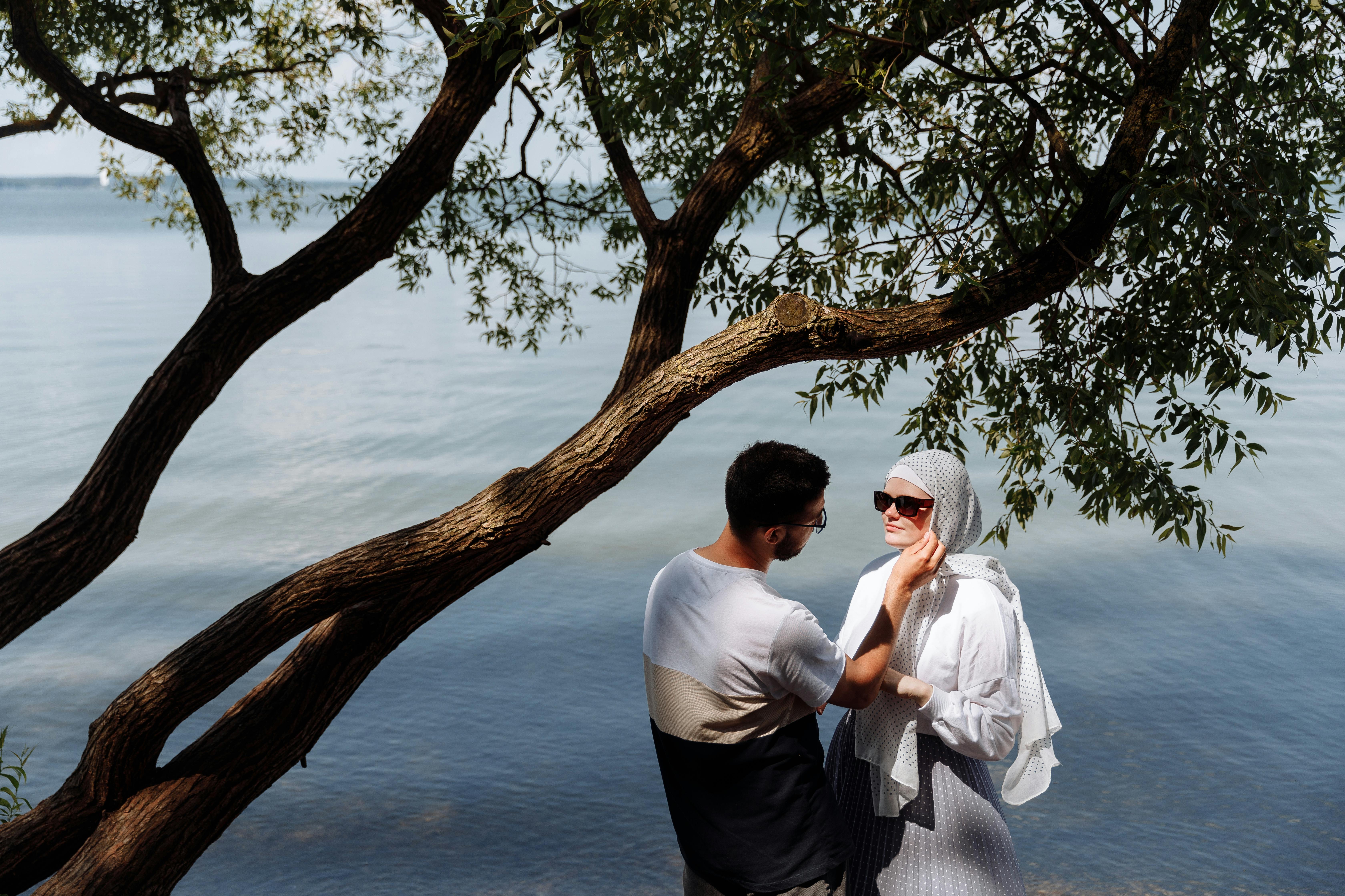 A loving couple enjoys a tender moment by a tranquil lakeside under leafy trees.