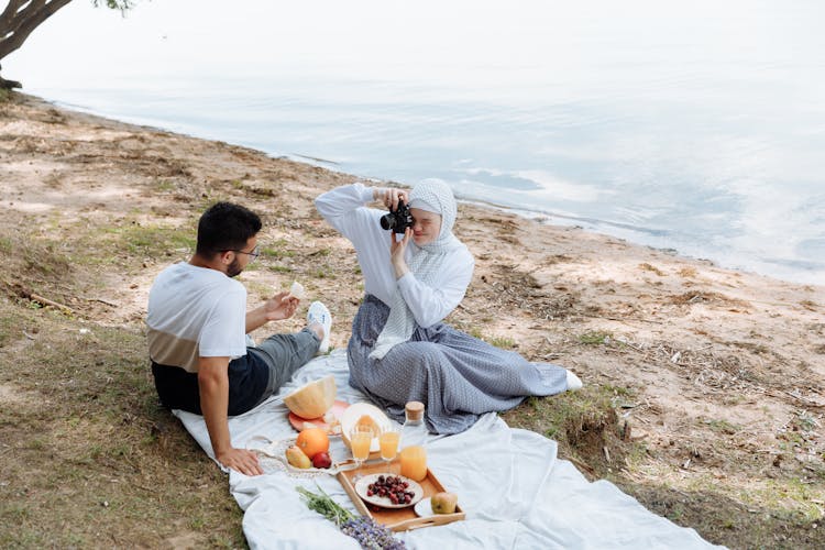 A Couple Having A Picnic Near The Water And Taking Photo