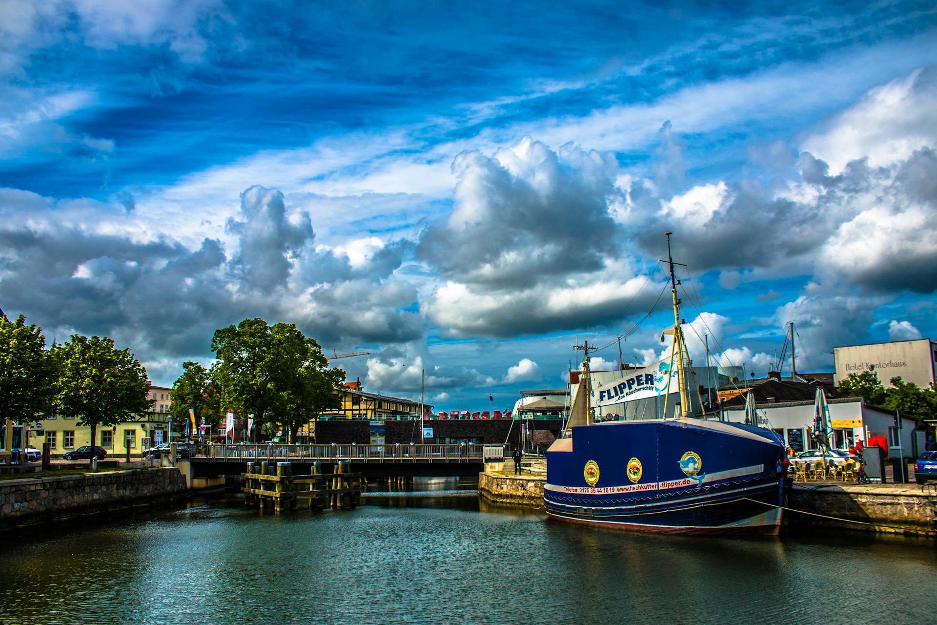 A vibrant view of Stralsund harbor featuring a sailboat, blue skies, and a bridge in the background.