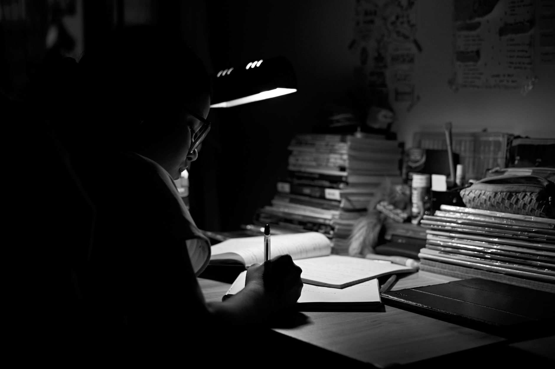 A student intensely studying under a desk lamp, surrounded by books, in a black and white setting.