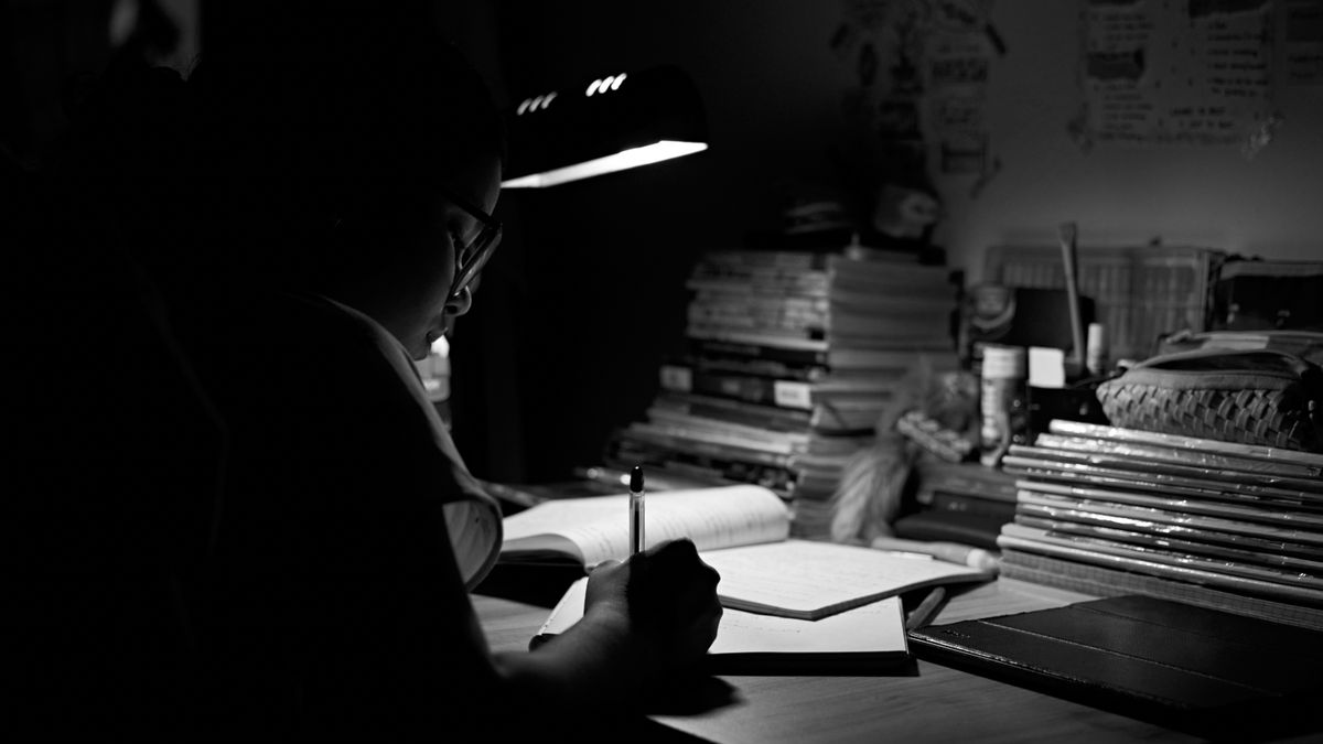Pessoa de óculos estudando e escrevendo em papel sob a luz de um abajur à noite