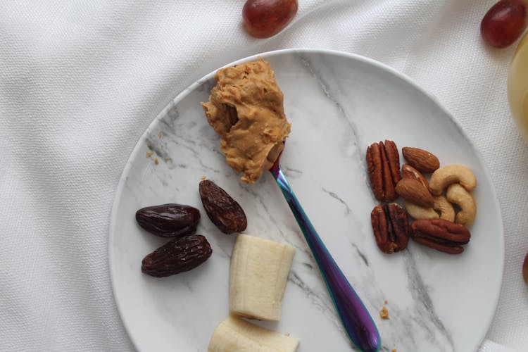 Nuts And Slices Of Banana On White Ceramic Plate With Spoon Of Peanut Butter