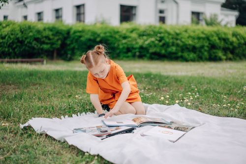Free A young girl in an orange dress reads books outdoors on a white blanket. Stock Photo