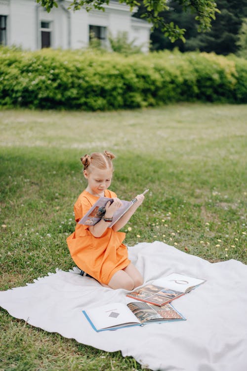 Girl In Pigtail Puffs and Orange Dress Reading A Book On Grass With A Picnic Blanket