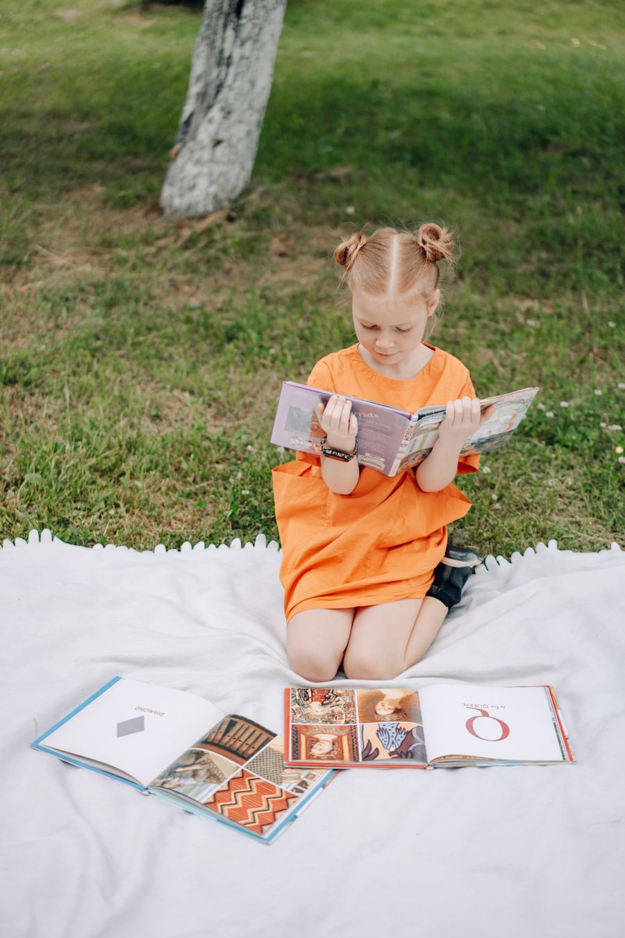 A young girl reading a book happily on a blanket outdoors