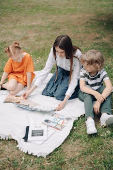 A teacher engages with two children reading a book on a picnic blanket outdoors.
