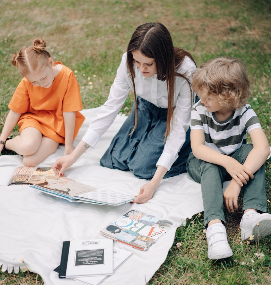 A teacher reading a book with two children on a blanket outdoors