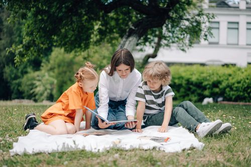 Free Three kids reading a book on a picnic blanket in a park under a tree. Stock Photo