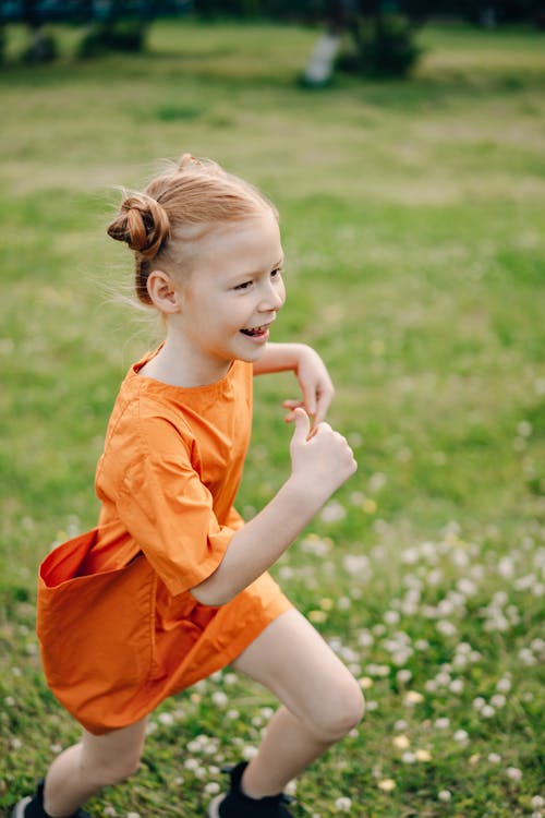 A Girl in Orange Dress Smiling while Running on Green Grass Field