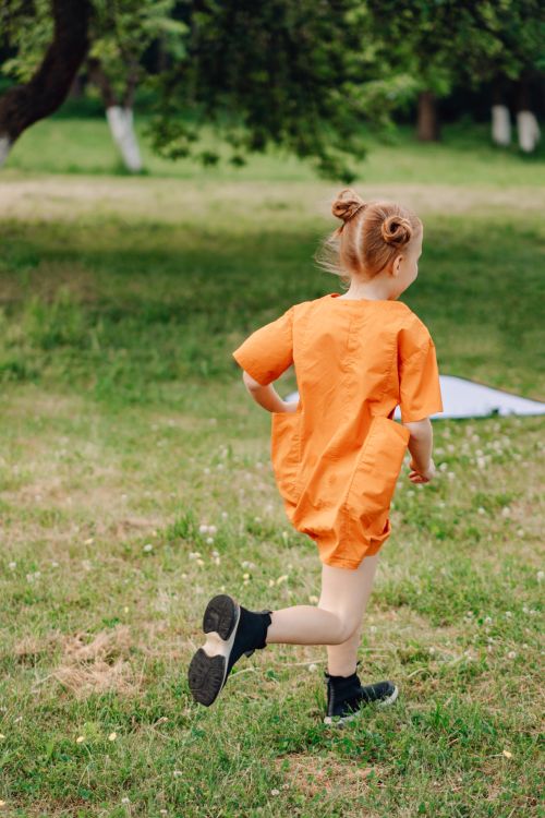 Free A joyful child with hair buns running through a grassy field in an orange dress, full of motion and energy. Stock Photo