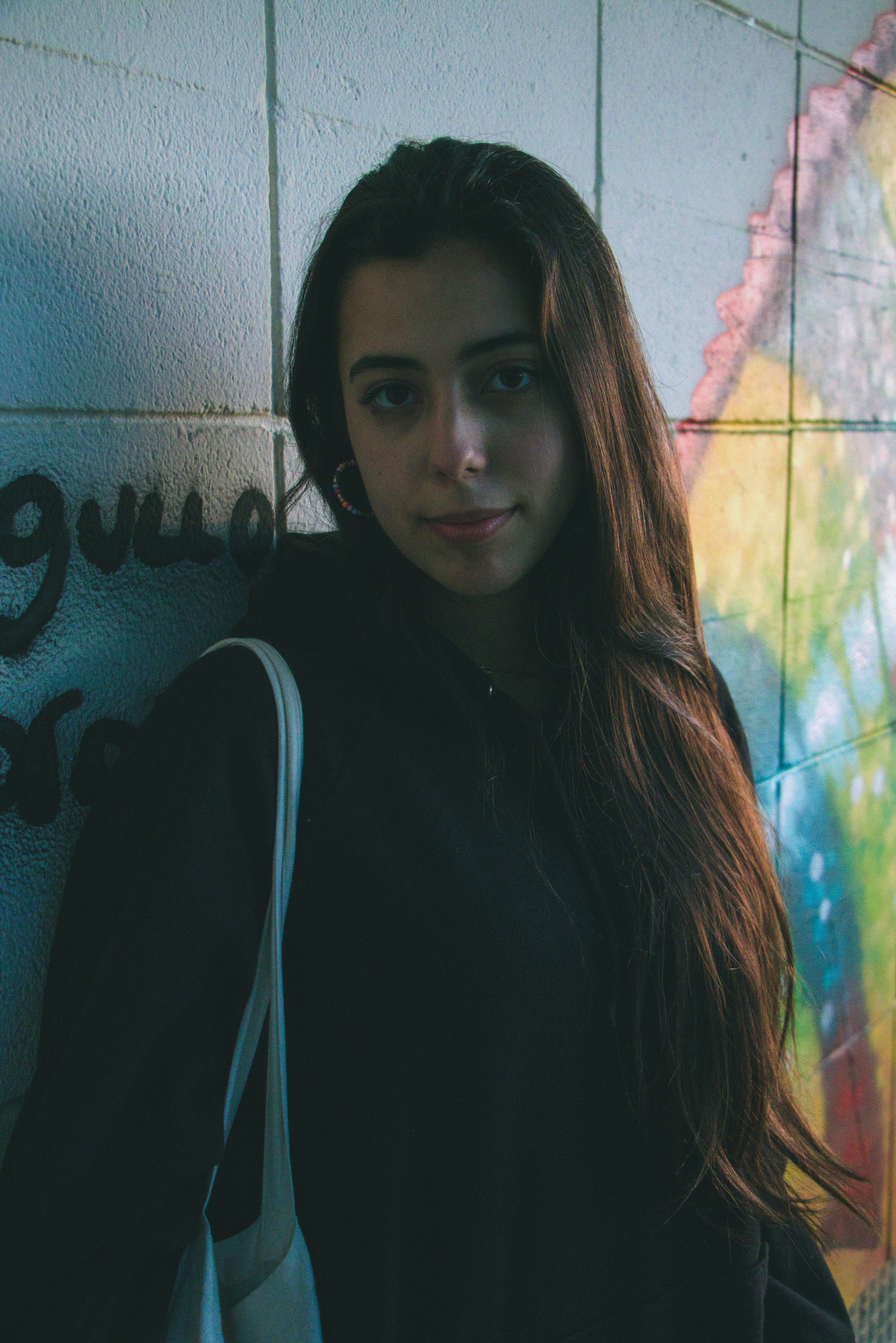 A woman in black long sleeves leans against a vibrant wall mural, smiling softly.