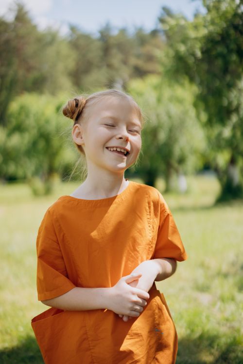 Free A joyful blonde girl in an orange dress enjoys a sunny day outdoors, smiling freely. Stock Photo