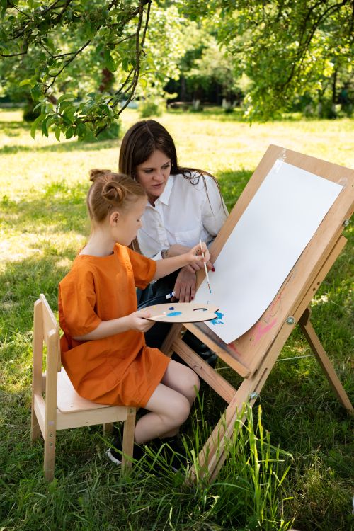 Free A child learning to paint with a teacher outdoors on a sunny day, capturing creativity and leisure. Stock Photo